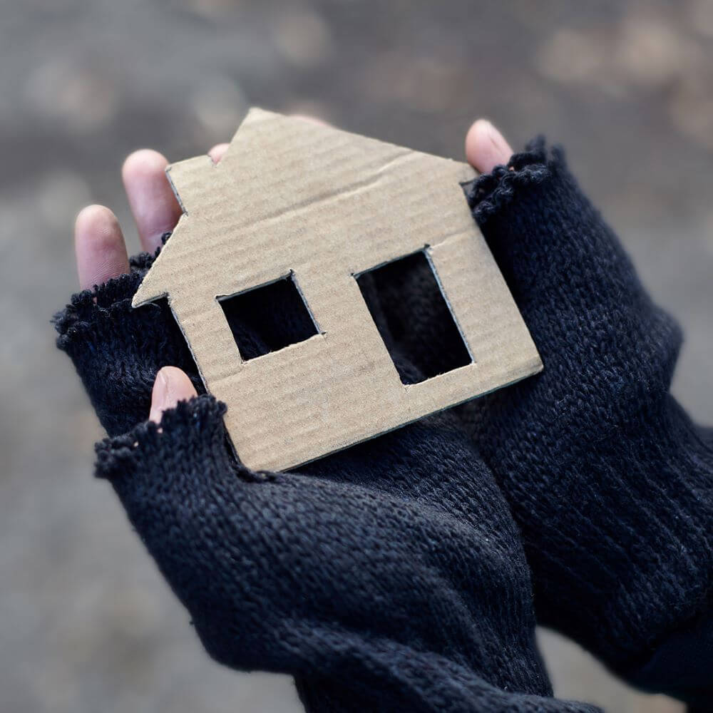 A hand wearing torn wool gloves holds a cardboard cutout shape of a house.