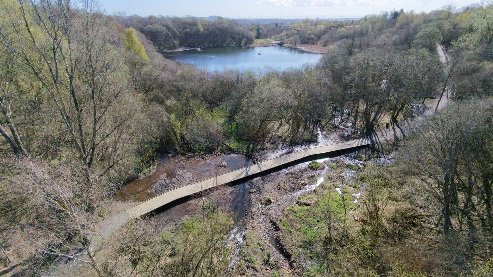 Boardwalk crossing a restored river channel and natural wetland area, with a pond and woodland in the background